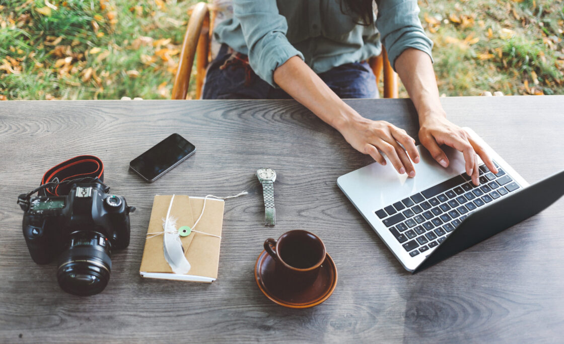 En contre-plongée, une femme écrit sur un ordinateur. Sur la table se trouvent un appareil photo, une carnet, une montre, un café et un téléphone portable.